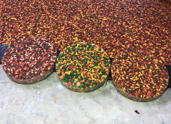 A photograph of three circular samples showing colours aggregates of RubbaSmart wet pour rubber in front of a pre-laid red, yellow and brown rubber surface. One sample is red, brown, and peach, the next is green, yellow and red, and the last is red, yellow and brown