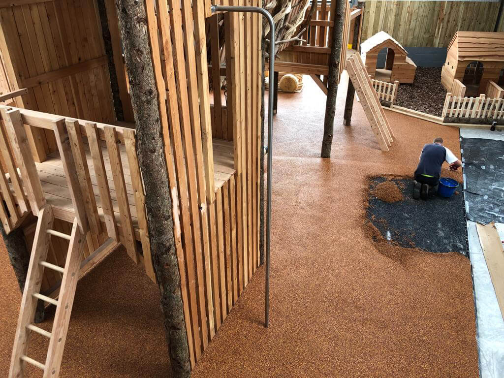 A photograph of one of Wet Pour Rubber installation. The brown rubber covers a playground surface, around wooden play structures and a workers is laying and smoothing out a final section