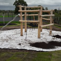 An image of RubbaSmart wet pour rubber playground surfacing being used on a playground. Over green artificial surfacing, blue wet pour is used to create a puddle shaped design beneath some playground equipment.